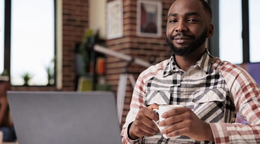 Portrait of confident freelance programmer looking at camera taking a break from coding holding a cup of coffee. African american student relaxing at home watching social media content on laptop.