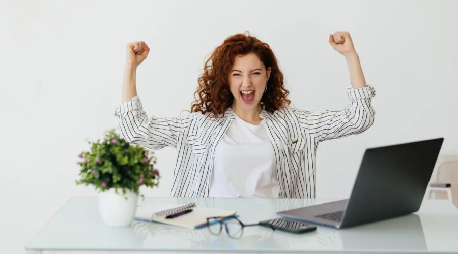 Young redhead woman working with her laptop raising fist after a victory, winner concept.